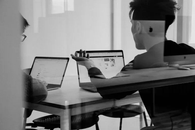 Two professionals in conference room have discussion while looking at laptops.