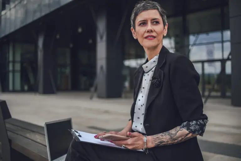 mature woman sits in front of office building with her laptop, notepad and phone in front of her